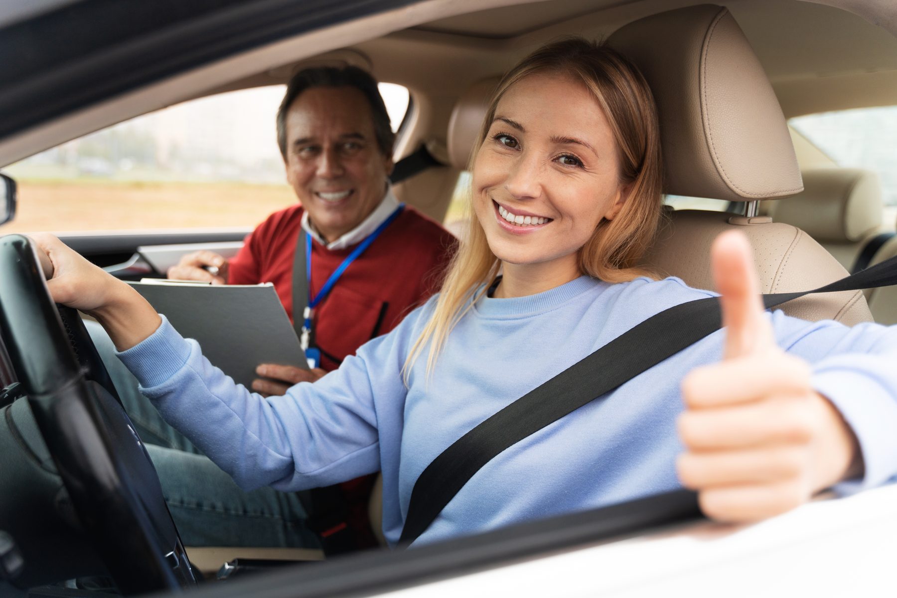 Learner driver giving a thumbs up at the wheel while her DVSA approved instructor reviews notes in the back seat